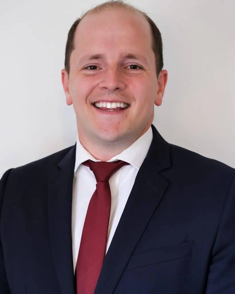 A professional headshot of a smiling man wearing a dark navy suit, white shirt, and burgundy tie. He has short hair and is standing against a plain light-colored background.