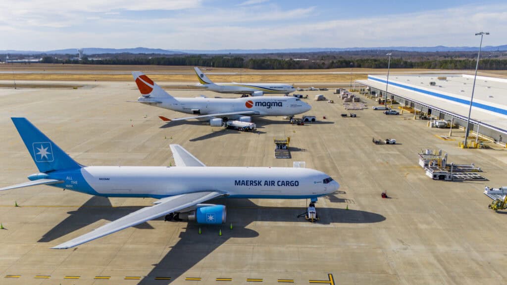 An aerial view of three cargo aircraft parked at an airport cargo apron near a warehouse facility. The nearest plane is a Maersk Air Cargo jet with blue and white livery, while the second features Magma Aviation branding, and the third aircraft in the distance Ground service equipment and cargo pallets are positioned nearby, with clear skies and tree-lined runways in the background.