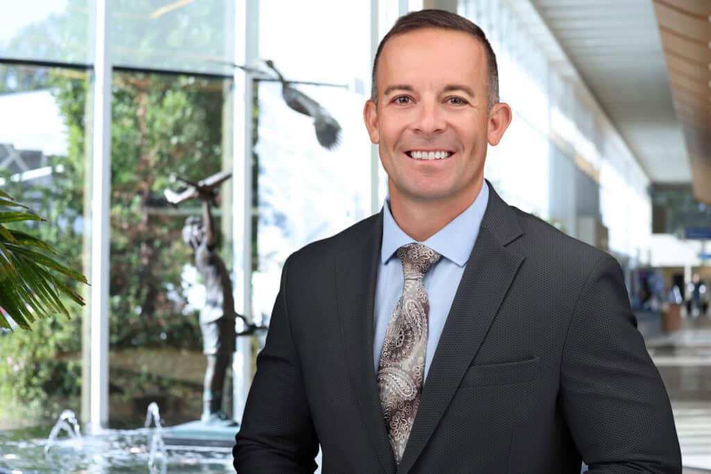 COO in dark suit with light blue shirt standing in front of fountain with bronze statue of a boy aviator holding an airplane.