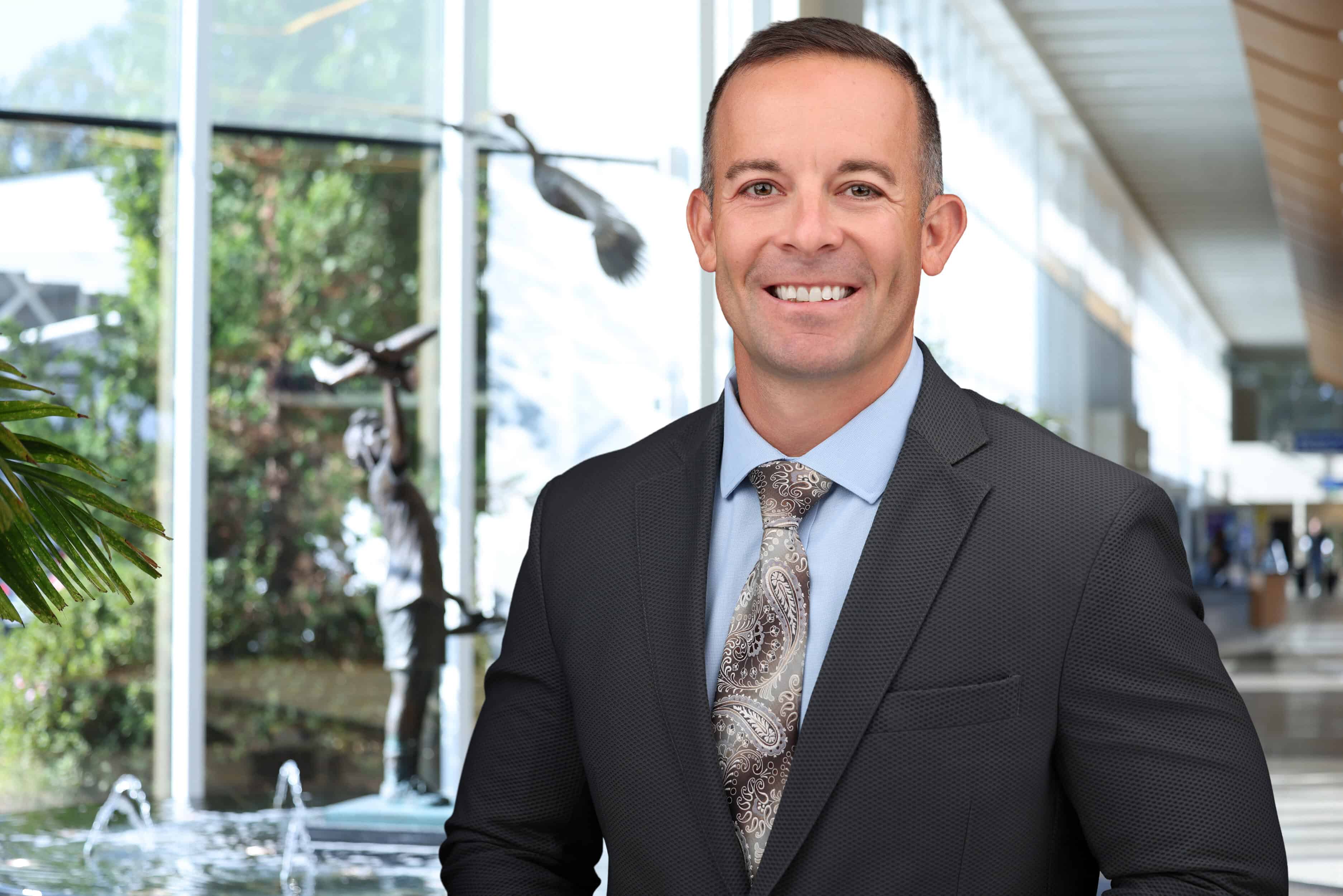 COO in dark suit with light blue shirt standing in front of fountain with bronze statue of a boy aviator holding an airplane.
