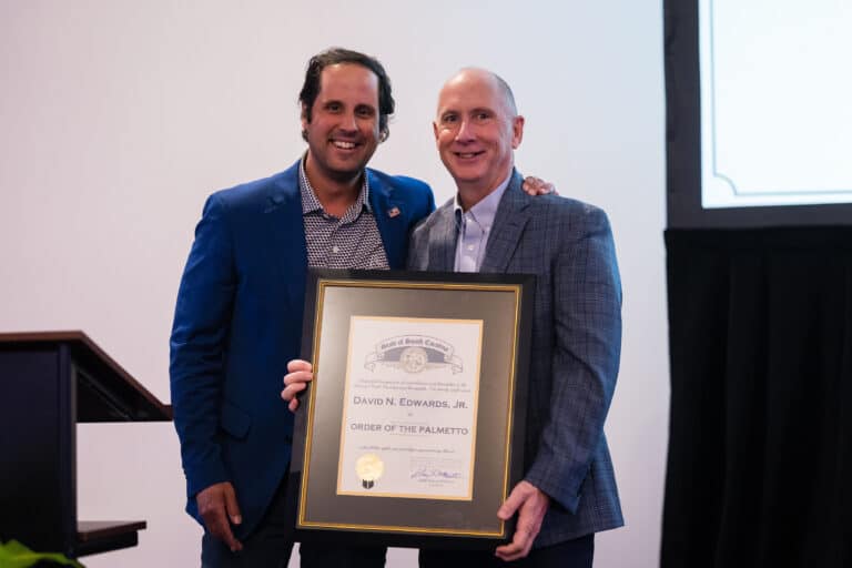 South Carolina Senator Josh Kimbrell stands beside Greenville-Spartanburg International Airport President and CEO Dave Edwards. Both are smiling as they hold a large framed Order of the Palmetto certificate presented to Edwards