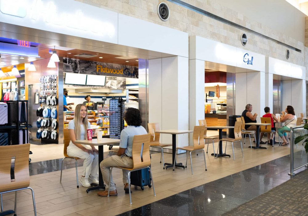 Passengers sitting at tables in front of Hudson, Flatwood Grill, and Dunkin' located on the first floor of the terminal near baggage claim.