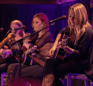 Autumn McEntire playing guitar in the foreground with Marti Dodson and SJ McDonald in the background.