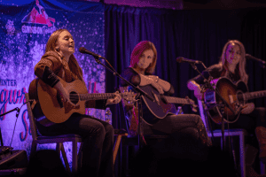 SJ McDonald singing in the foreground with Marti Dodson and Autumn McEntire listening in the background.