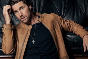 Country Music singer, John King, leans against a black leather couch wearing a black tee shirt and brown leather jacket.