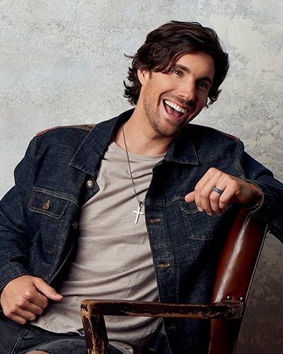John King laughing and leaning against a leather chair on one arm wearing a jean jacket, gray t-shirt, and jeans.
