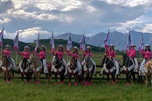 The Latigos Up Drill Team wearing matching pink uniforms performs at a Gunnison Tough Enough to Wear Pink event on horseback with flags.