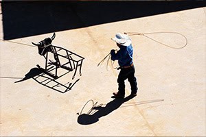 A little buckeroo in a cowboy hat and boots practices roping on a metal steer.