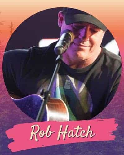 Rob Hatch playing a guitar wearing a black tee and baseball cap in front of a microphonr.