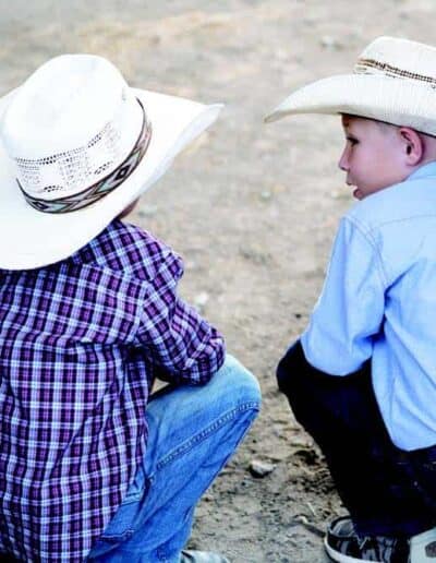 Two young cowboys with straw cowboy hats kneel in the arena and talk.