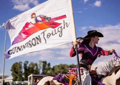 The Gunnison Tough flag waves brightly as a cowgirl in purple carries it on horseback.