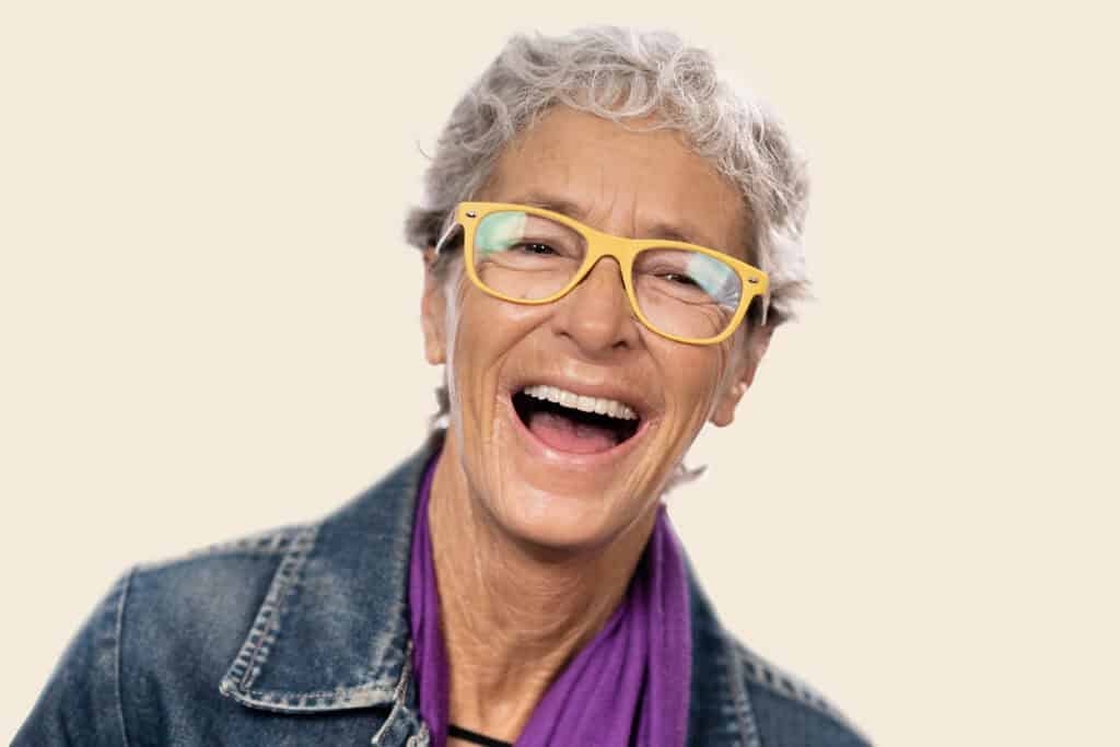 Senior woman with dentures smiling, wearing yellow glasses and a denim jacket.