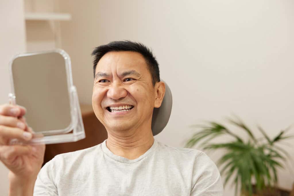 Smiling man examining his new full denture set in mirror.
