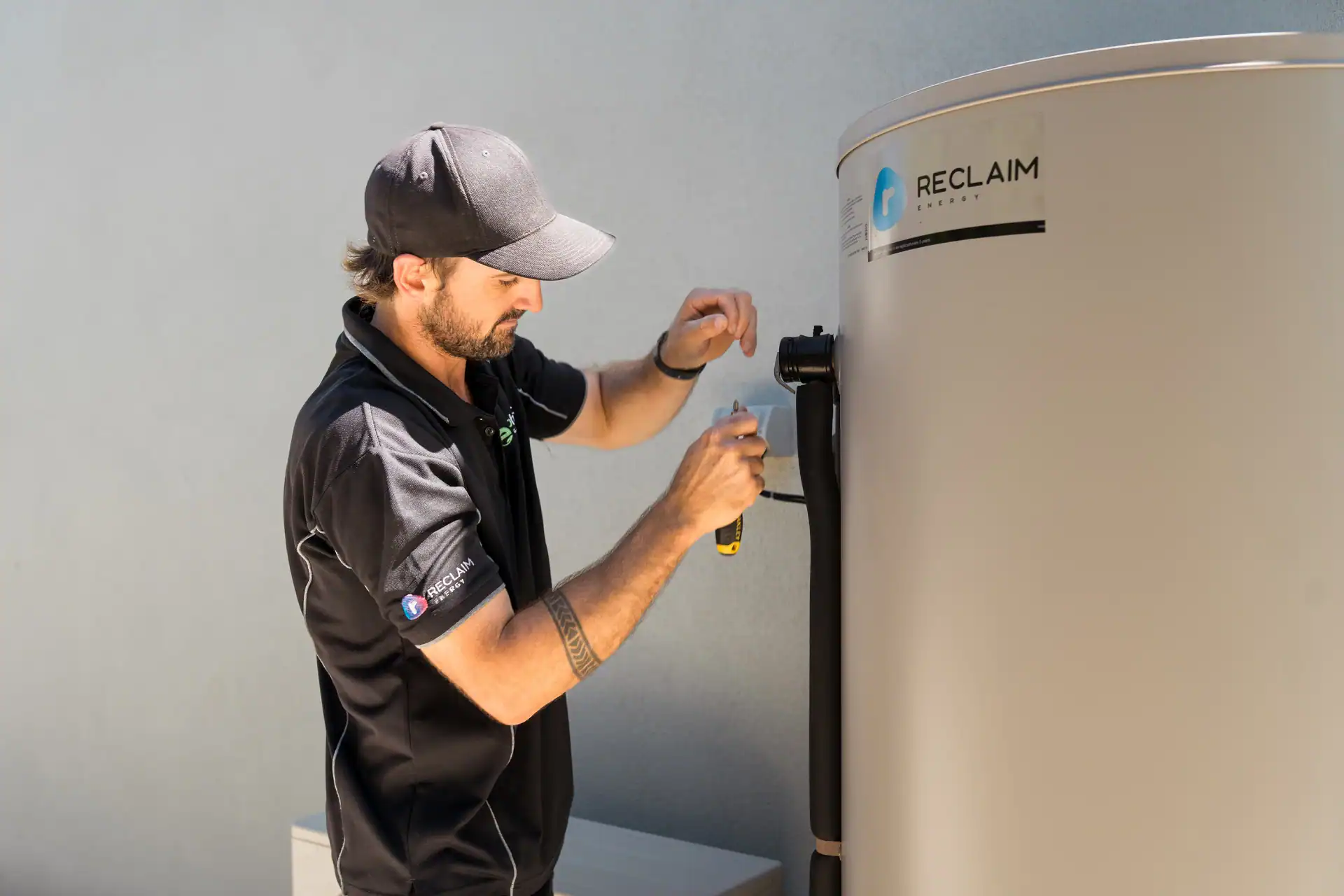 A technician in a black uniform uses a tool to service or install a Reclaim brand water heater against a light-colored wall.