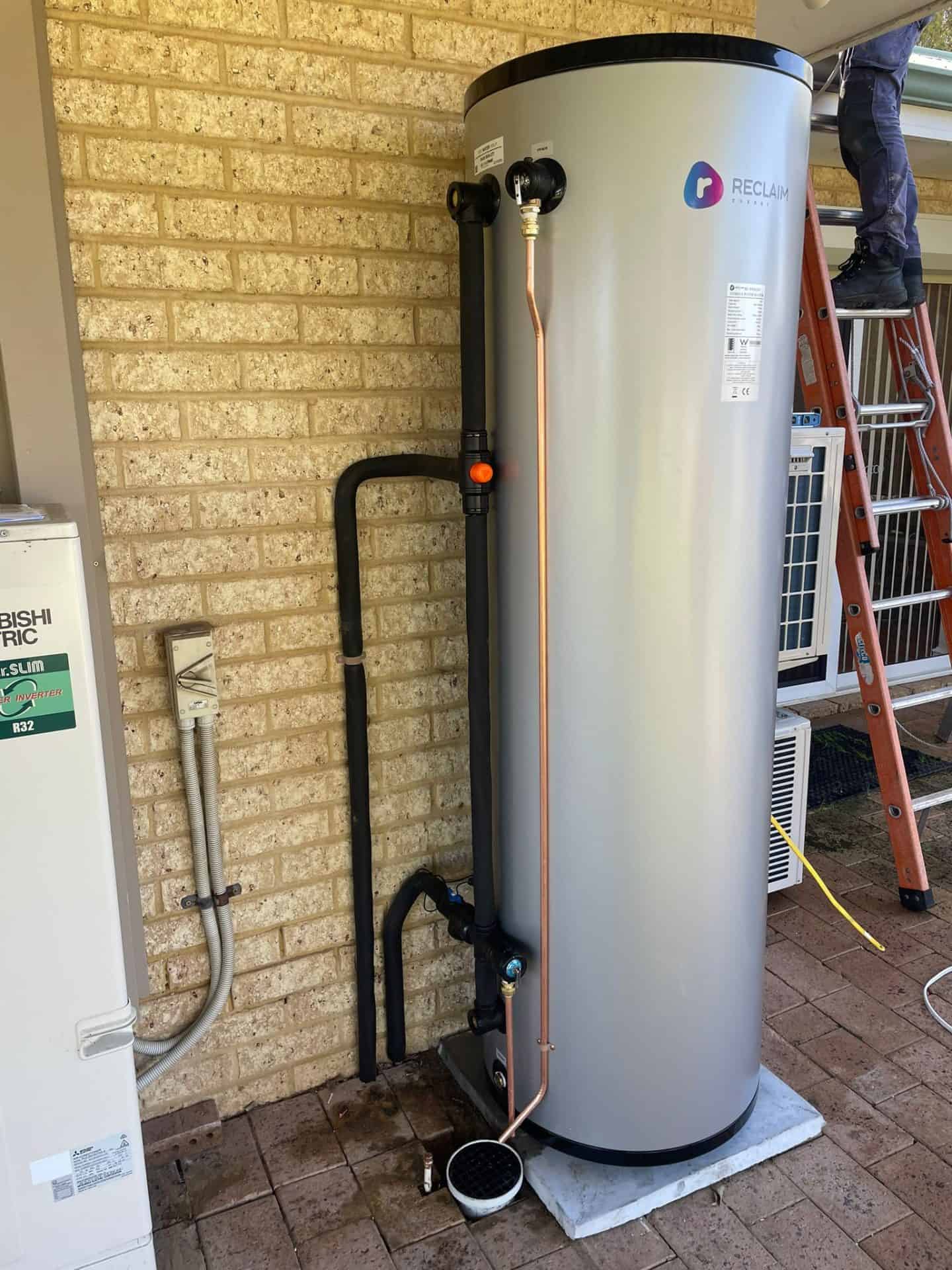 A tall cylindrical hot water system with pipes connected stands on a brick patio against a brick wall, near a ladder and air conditioning units.