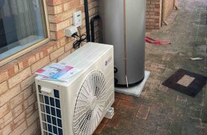 A heat pump hot water system installed outside a brick house, with an outdoor compressor unit and a tall cylindrical water tank.