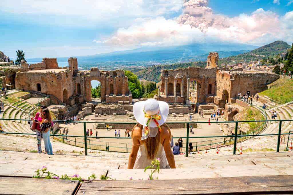 Girl with white hat looking to Ancient Greek theatre of Taormina, Sicily with Mt Etna erupting in the far distance.