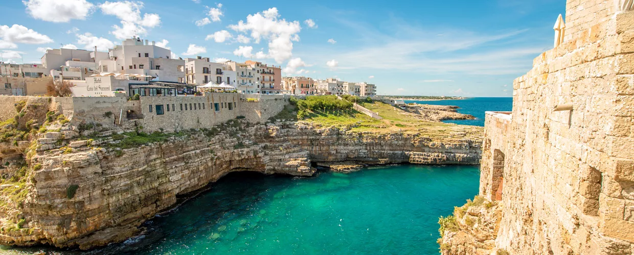 Aerial view of white stone buildings on a clifftop, on the seas edge, with turquoise water on a sunny day.