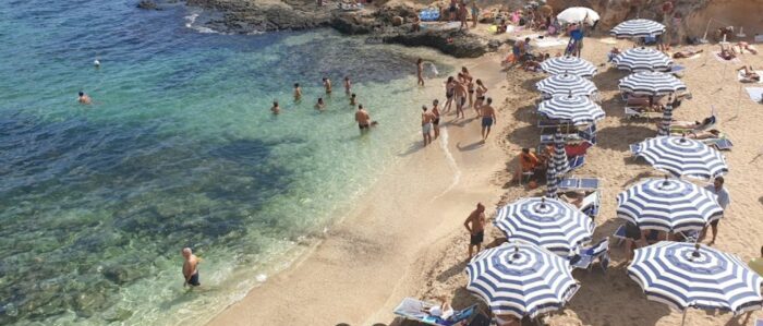 small sand beach with people bathing in shallow clear sea and others on the beach with blue and white stripoed beach umbrellas and deck chairs