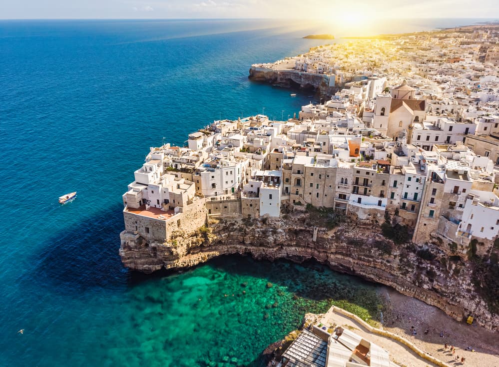 Aerial view of pastel coloured, densely built houses on clifftop and Lama Monachiles narrow beach nestled between the high cliffs, with turquoise coloured sea on a sunny day.