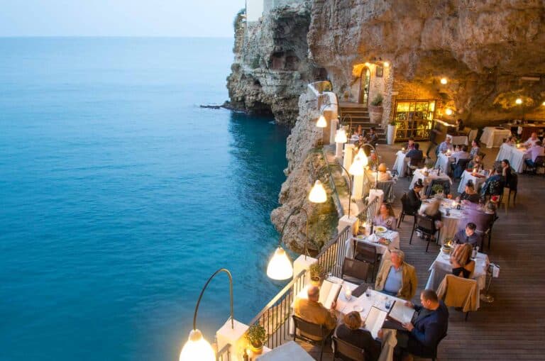 people dining at the edge of the sea cliff in a cave restaurant overlooking the blue sea at dusk.