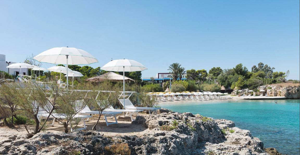 surrounded by rocks and vegetation with white sand beach and white beach umbrellas and deck chairs on a beautiful sunny day