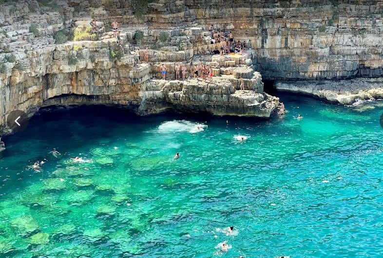 people swimming in turquoise sea with rocky cliffs in background and other people sunbathing on a rock ledge