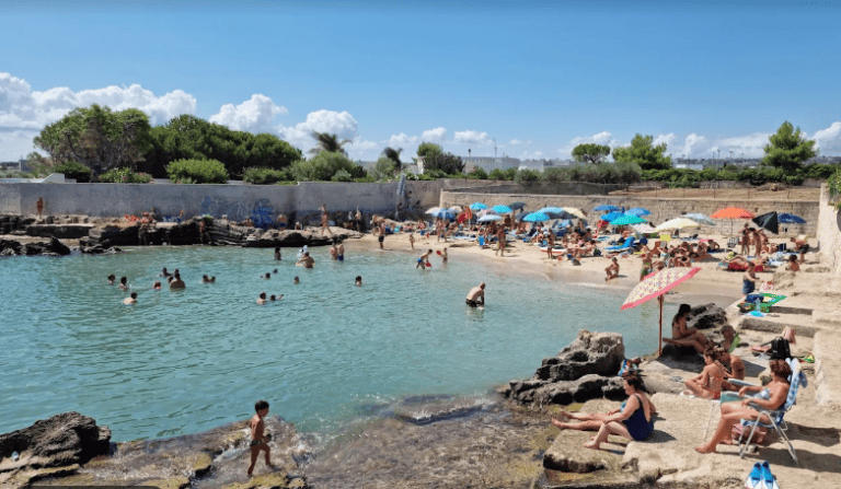 view of a white sand beach with people under beach umbrellas enjoying a beautiful summers day,  and people in the shallow water bathing. 