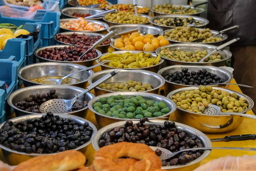 close up image of stainless steel bowls filled with black and  green olives in various shades on a market stall bench.