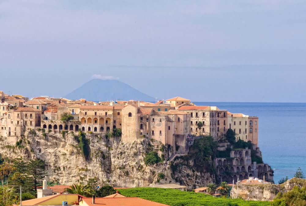 Aerial view of medieval period buildings 2 to 3 storeys high resting on high cliffs, with the sea and a silhouette of a cone shaped island in the distance known as Stromboli.