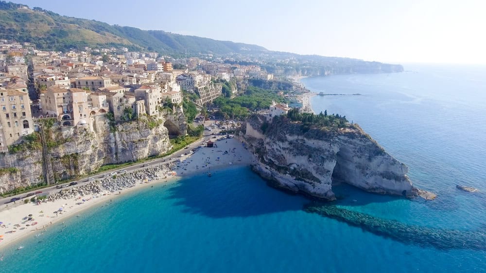 Aerial view from a drone over the blue sea looking towards old buildings of Tropea sitting on the cliffs and a white sand beach below, 