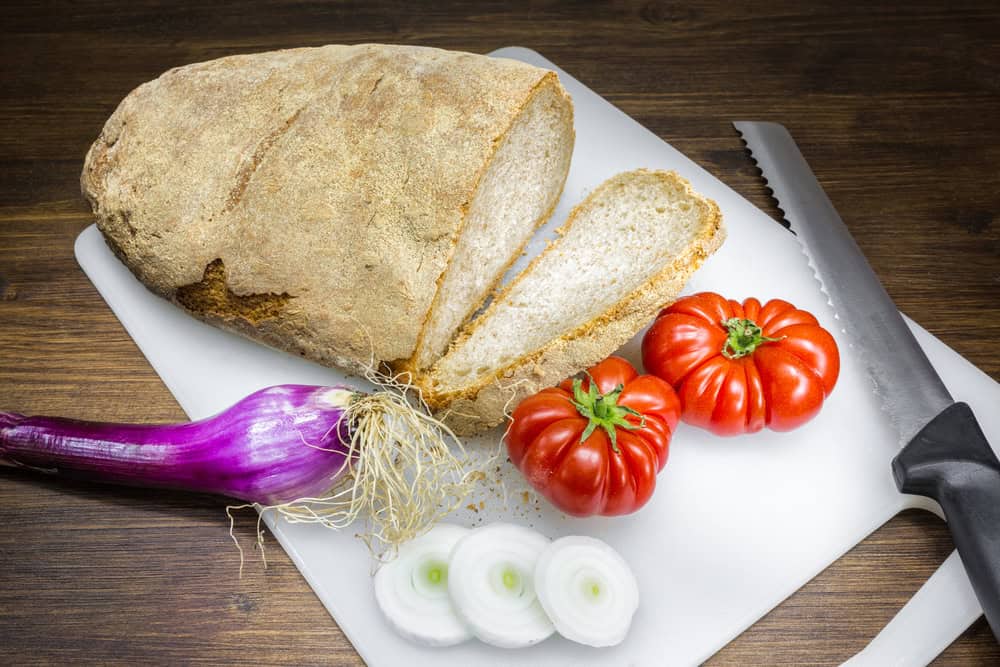 Wood fired light brown bread loaf with slices cut and purple onion of Tropea sliced and 2 red tomatoes on a plastic cutting board.