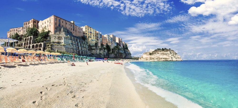 view from the turquoise blue waters edge of a white sand beach of Tropea looking back towards old pastel coloured buildings on the cliff edge and a rocky outrop with a church on top in the far distance.