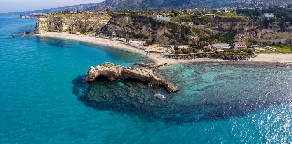 Aerial view from a blue sea on a sunny day  looking back to land and a white sand beach and rocky outrcop stretching out seaward of Baia di Riaci and its beachfront cafe and restaurant in the background at the foot of the cliffs behind