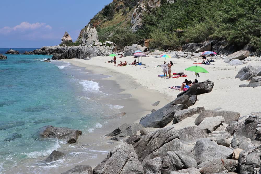 Michelino, a secluded white sand beach with outlying rock outcops and blue sea on a sunny day. several coloured beach umbrellas and some people strolling on the waters edge. A cone shaped rock in the distance called the Pizzuta.