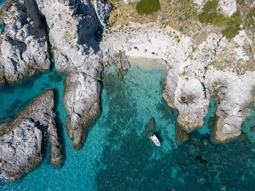 aerial drone view of a rocky coastline and a secluded small white sand beach with 3 beach umbrellas and one person swimming,  near capo vaticano on a sunny day and a single boat floating on a blue transparent sea.