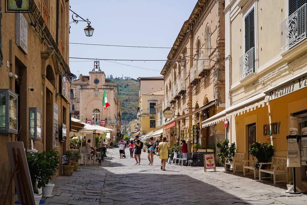 cobble stoned street of Tropea in old town with olkd stone buildings 2 to 3 storys high on each side. chairs and table on the street from cafes and people strolling. Also a stone church  in the distance.