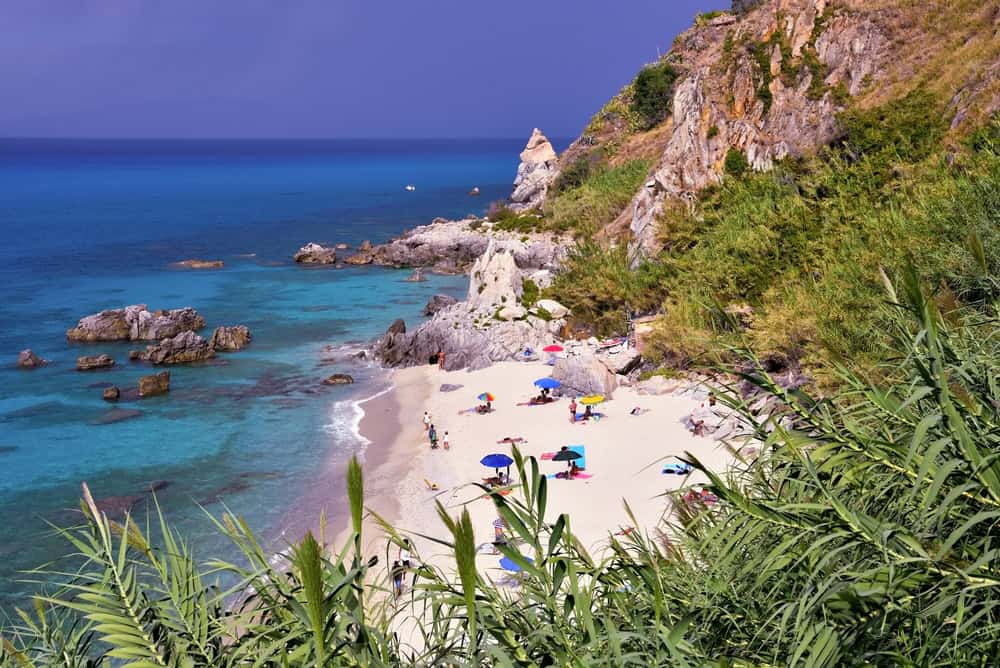 Michelino, a secluded white sand beach with outlying rock outcops and blue sea on a sunny day. several coloured beach umbrellas and some people strolling on the waters edge.