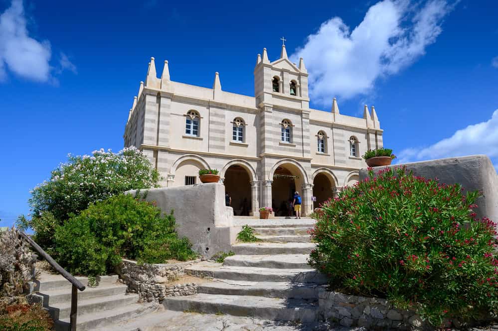 Close up of a stone church with 3 arches at ground level and smaller arched windows on the upper level. Stone steps leading up to the entrance and green plants either side and a clear blue sky with some puffy clouds.