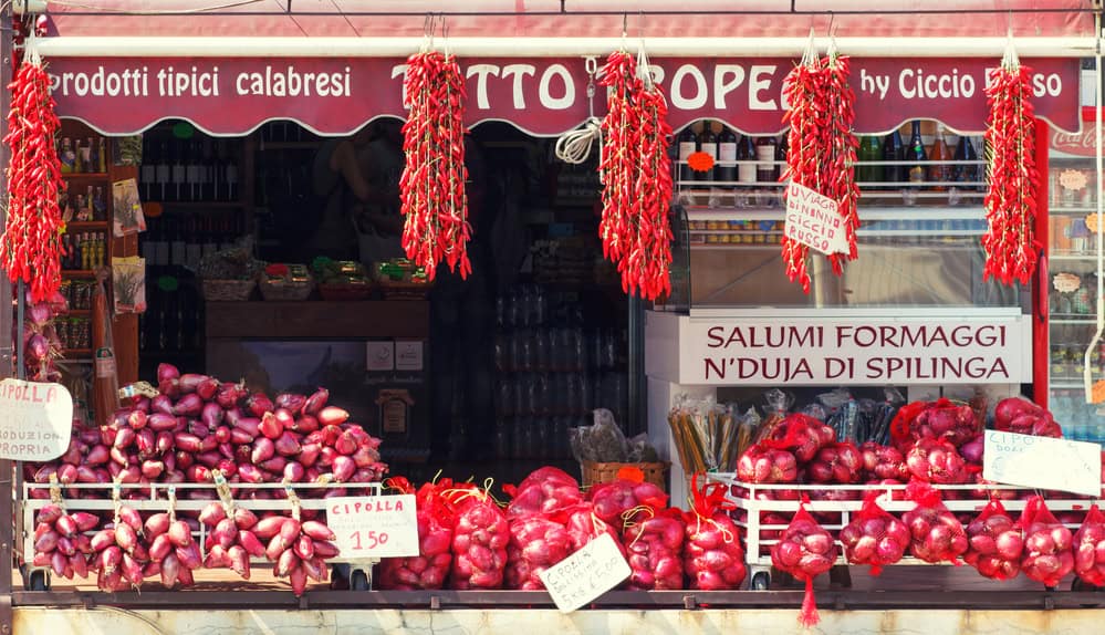 small shop with red awning over, with strings of fresh chillies hanging and bags of red tropea onions on a stall at the front of shop. Sign that says typical calabrian products.