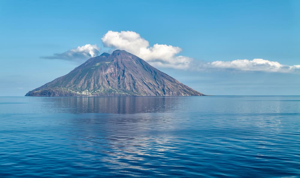 close up view of a cone shaped island stromboli from a calm blue sea with puffs of smoke and clouds above on a sunny day.
