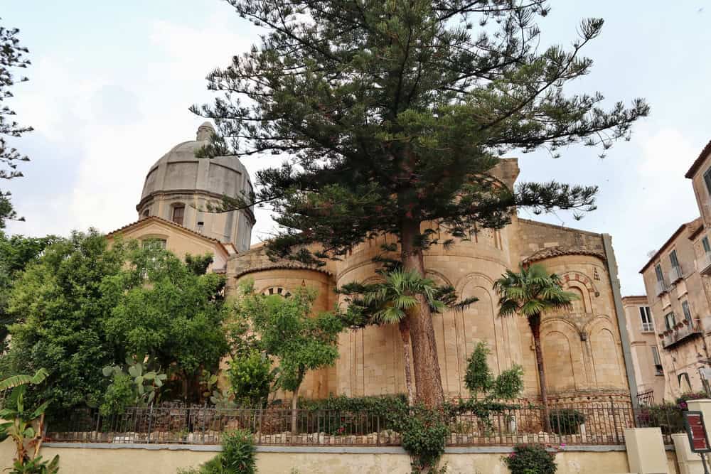 12th century stone cathedral of Tropea with curved walls and frescoes with a dome shaped bell tower behind . large green tree in front and smaller bushes on each side