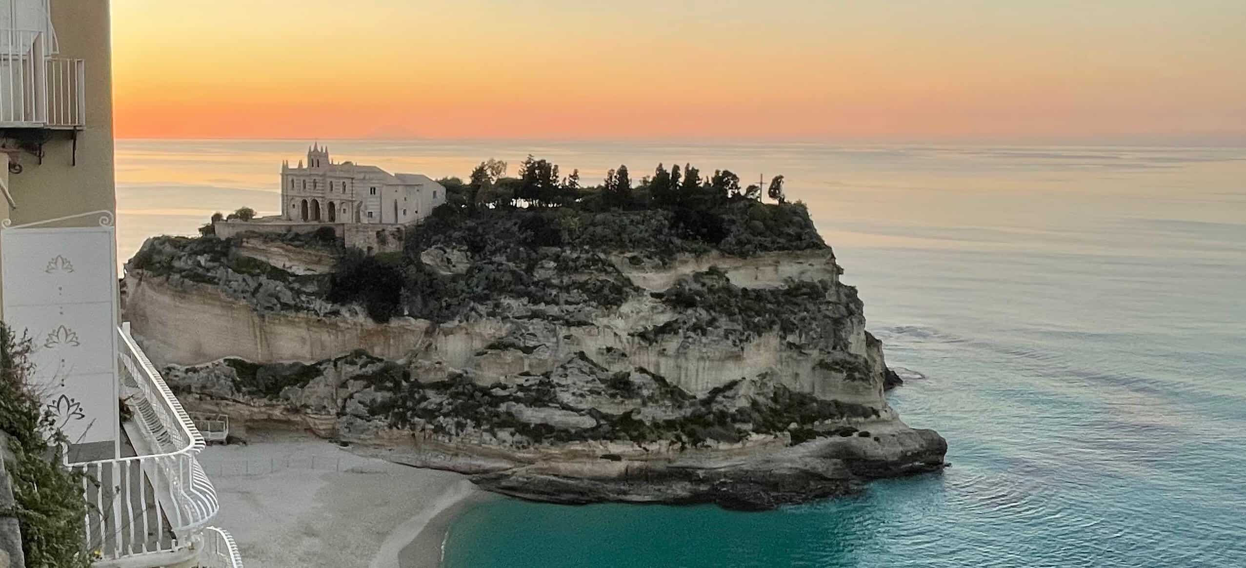 Historic church of Santa Maria della Isola in Tropea on rocky outcrop with white sand beach and colourful beach umbrellas.