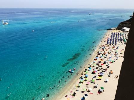  white sand beach and blue sea in foreground, and sun umbrellas and bathers in the water below. 