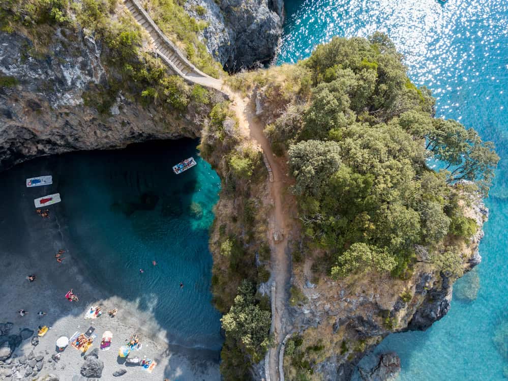 aerial image of a small secluded beach cove with the rocky outcrop above the arch and the path that leads down to the beach on a sunny day. A few people lying on towels and some beach umbrellas.