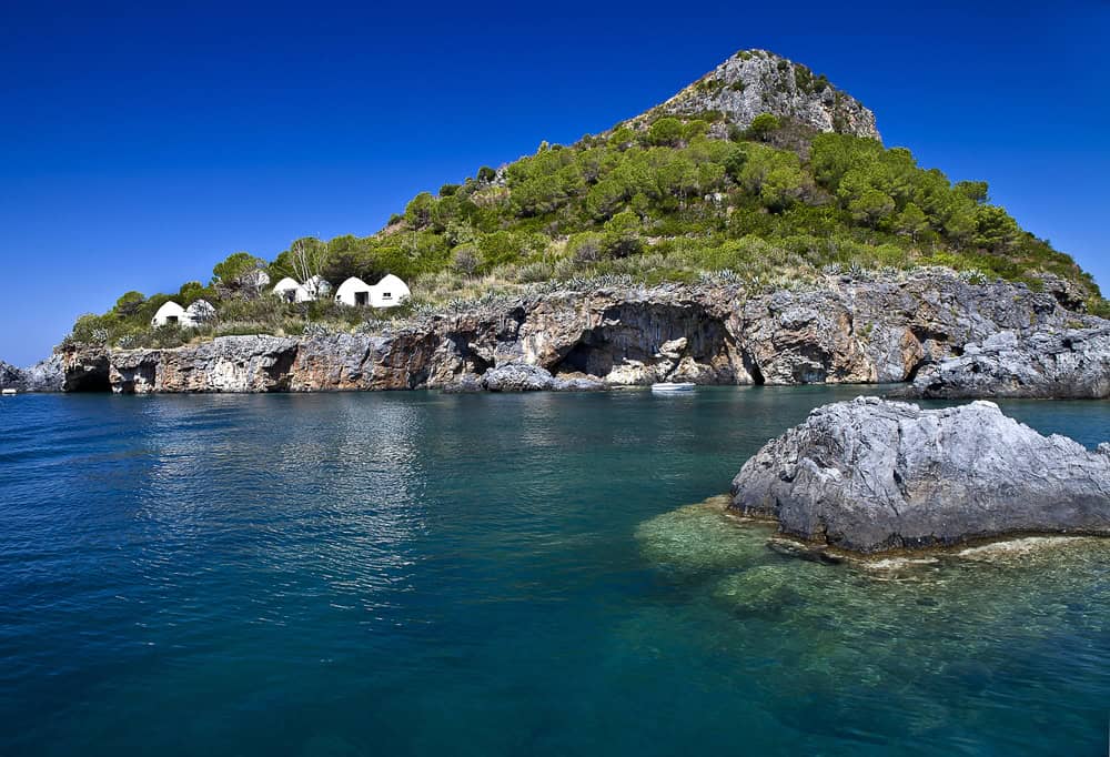 water level image towards a rocky Dino Island and some green vegetation with white painted dome shaped buildings at the top of the small cliff at the waters edge on a beautiful sunny day