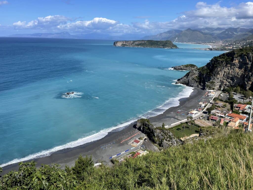 aerial view of a dark sand beach with turquoise sea and outlying island of Dino in the distance and mountainous coastline with green vegetation above on a sunny day