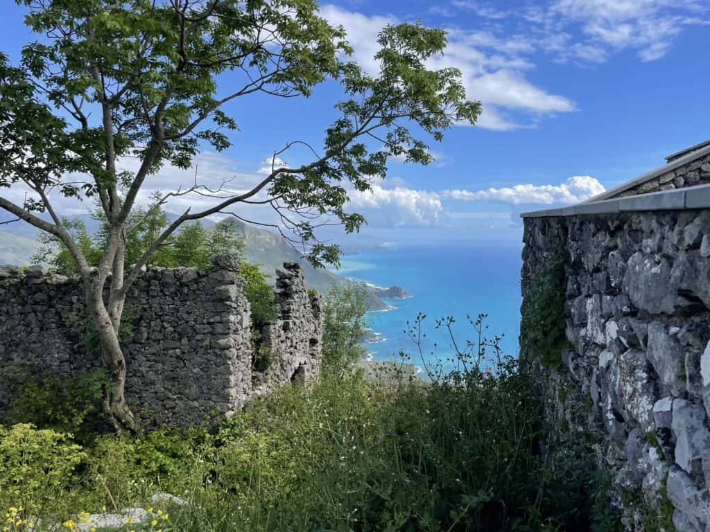 aerial view of the rugged coastline and blue sea in the distance and in the close foreground stone ruins of a building and a small tree nearby on a partly cloudy sunny day