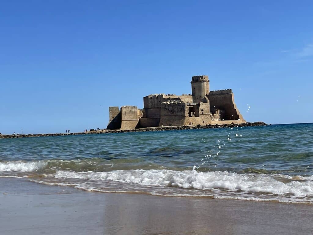 Seaside view of a stone castle and fortress appearing to float on the blue sea on a sunny day with small waves breaking on the shoreline 