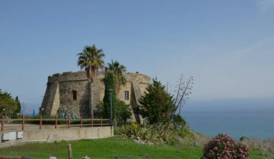 historic stone circular fortress on a hill with blue sea beyond ans 2 palm trees at the front of the fortress on a sunny day
